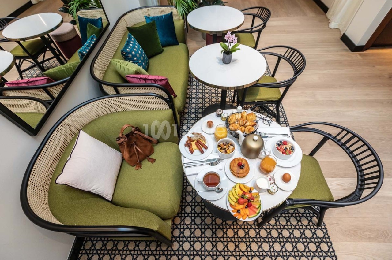 Table basse dressée pour un petit-déjeuner avec viennoiseries, fruits et boissons, entourée de fauteuils verts.