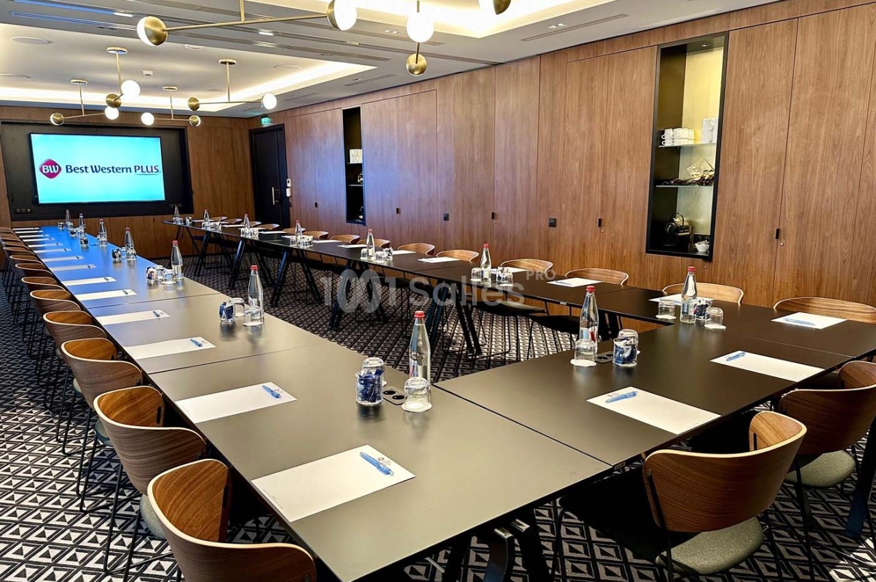 Salle de réunion moderne avec tables disposées en U, chaises en bois, bouteilles d'eau et écran affichant ’Best Western Plus…
