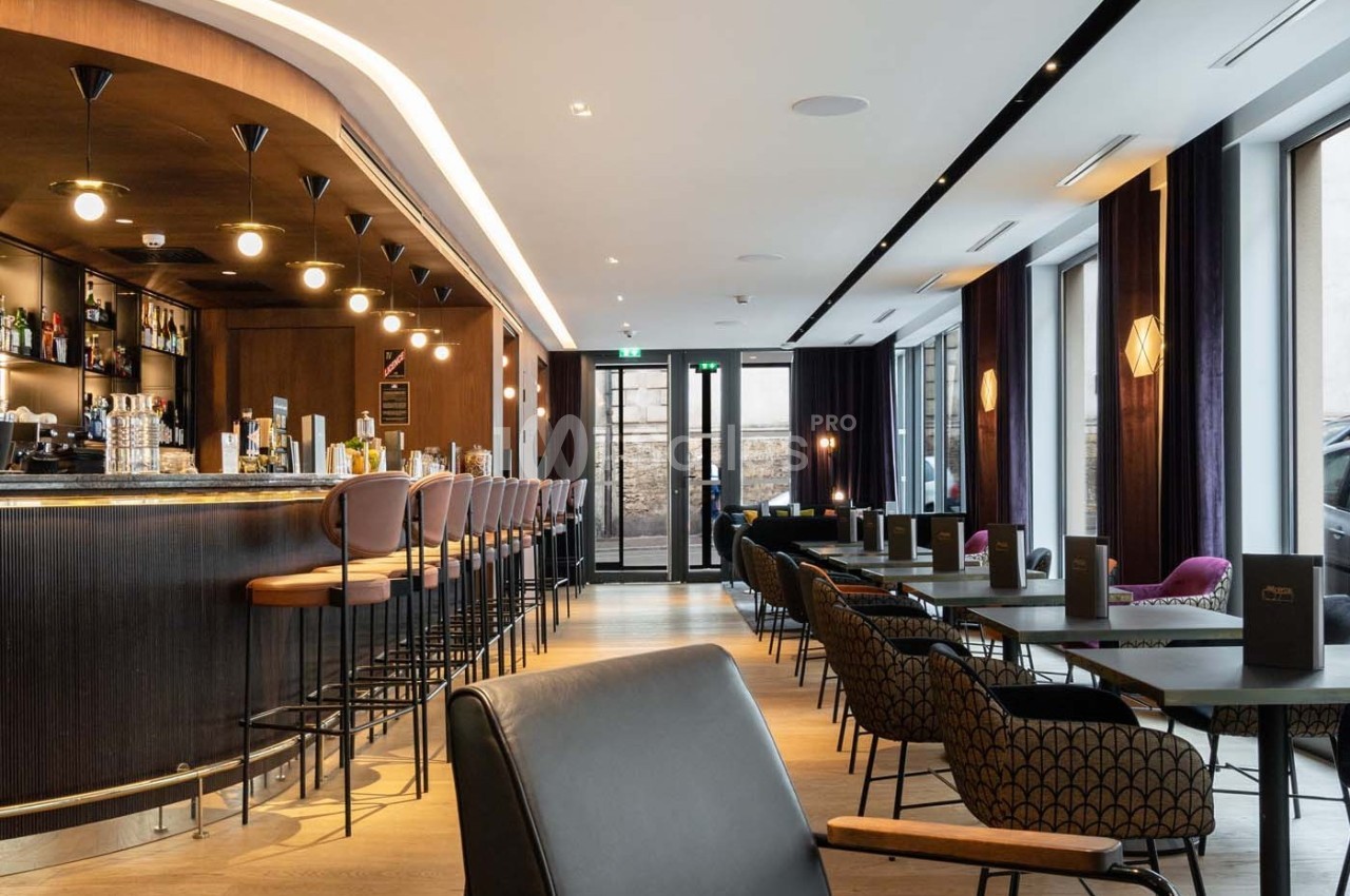 Bar moderne avec comptoir en bois, tabourets hauts, tables et chaises élégantes dans un espace lumineux et épuré.