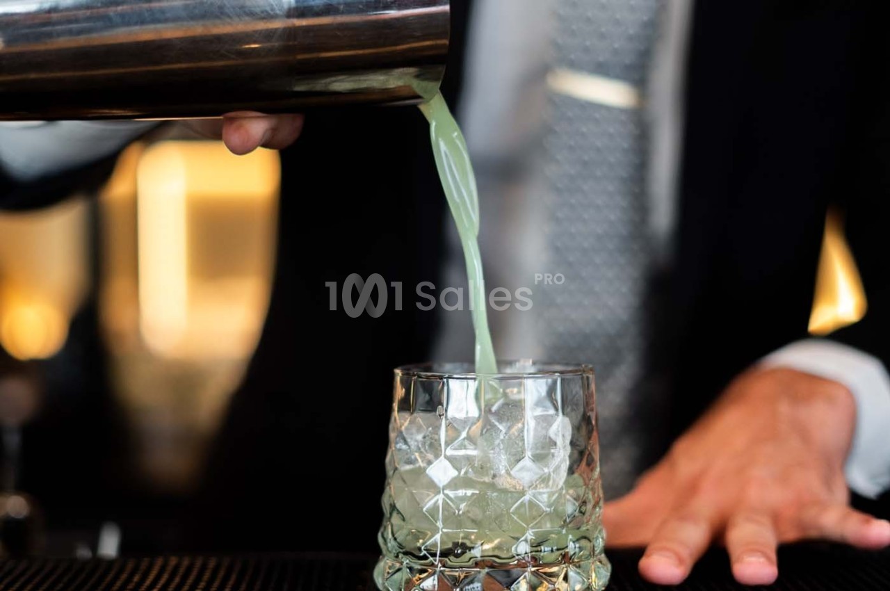 Un barman verse un cocktail vert dans un verre en cristal sur un comptoir.