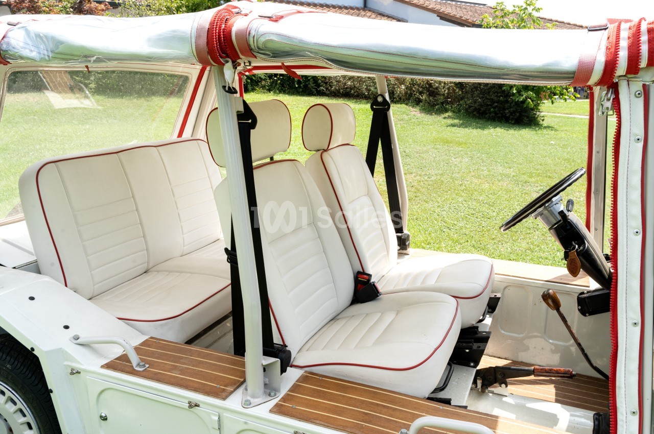 Intérieur d'une voiture décapotable vintage avec sièges blancs bordés de rouge, vue sur un jardin en arrière-plan.
