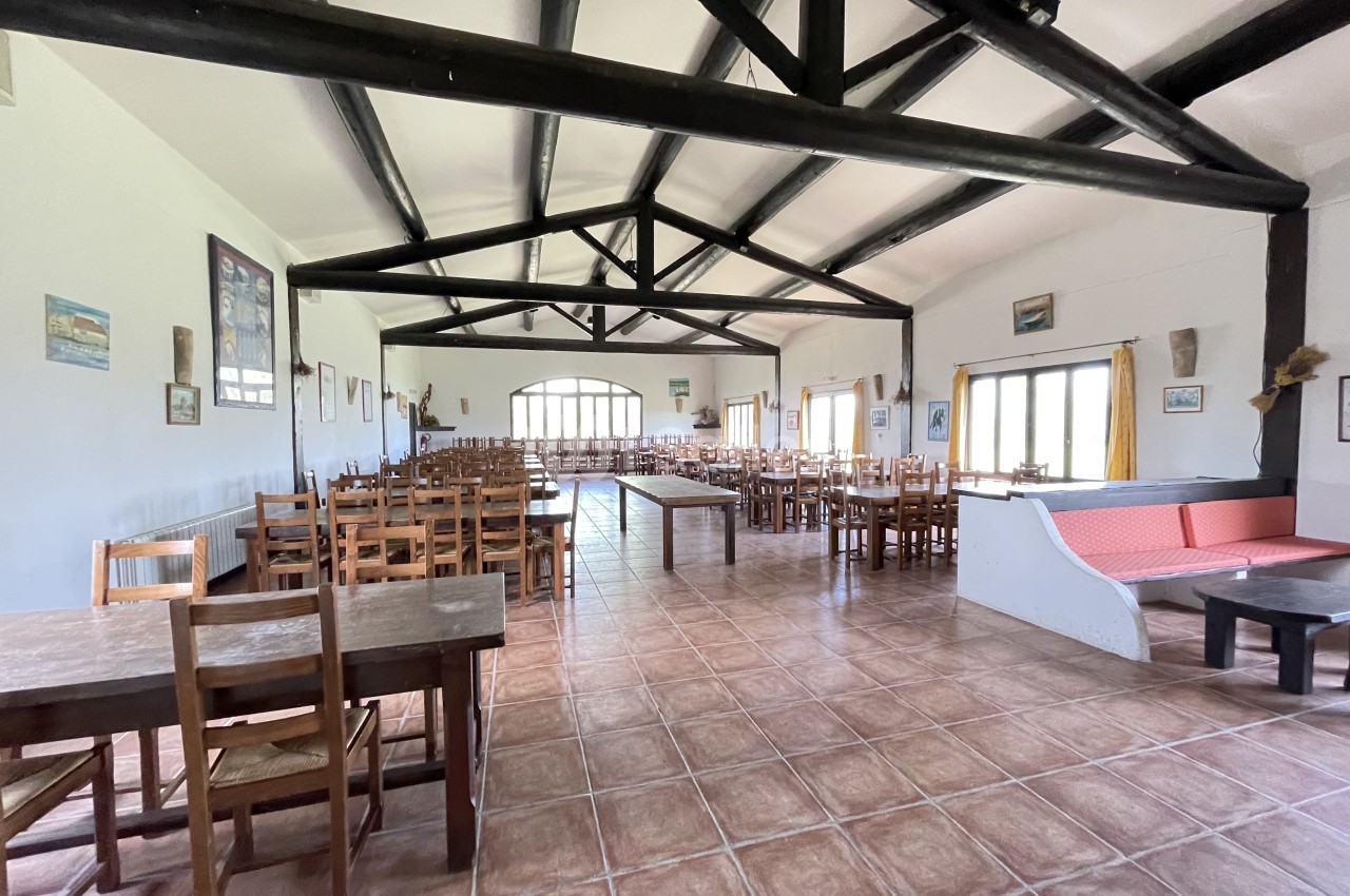 Salle spacieuse avec des tables et des chaises en bois, poutres apparentes au plafond et grandes fenêtres lumineuses.
