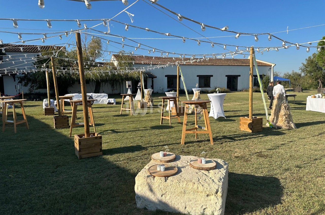 Espace extérieur aménagé pour un événement avec tables hautes, guirlandes lumineuses et bâtiment en arrière-plan.