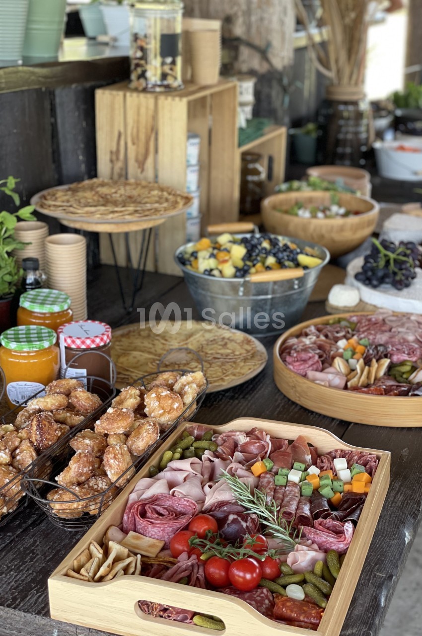 Plateau de charcuterie, fruits, légumes et condiments sur une table en bois avec d'autres plats et boissons en arrière-plan.