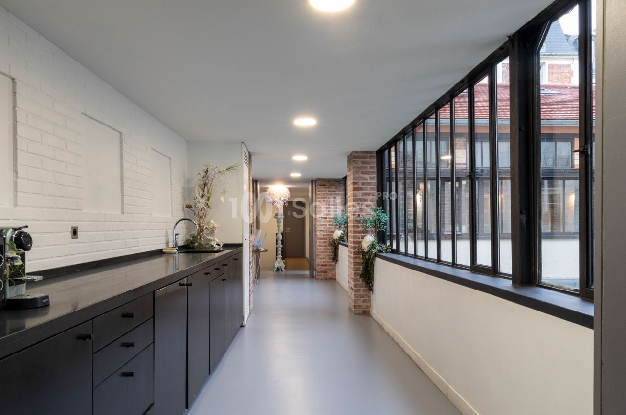 Cuisine lumineuse avec plan de travail noir, mur en briques blanches et grandes fenêtres donnant sur une cour.
