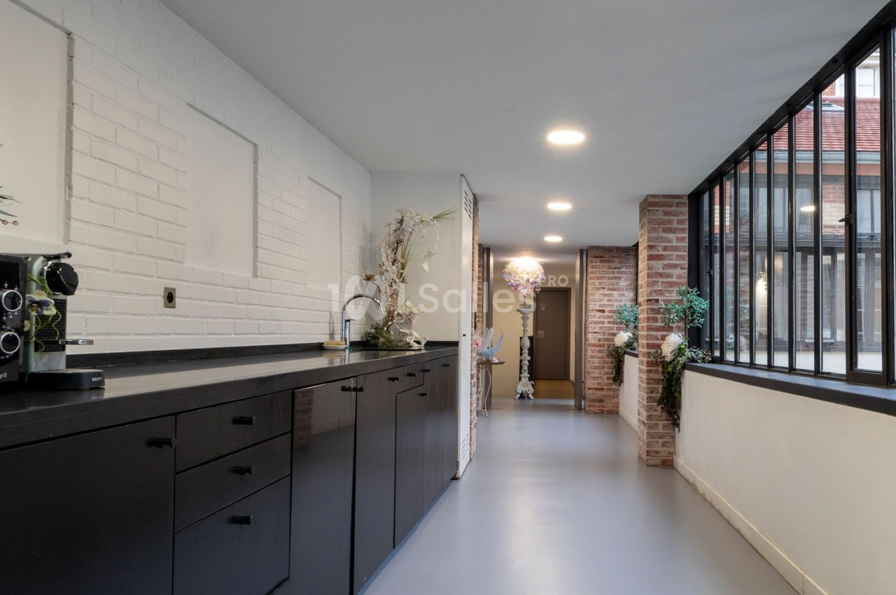 Cuisine moderne avec plan de travail noir, mur en briques blanches, grandes fenêtres et vue sur un couloir lumineux.