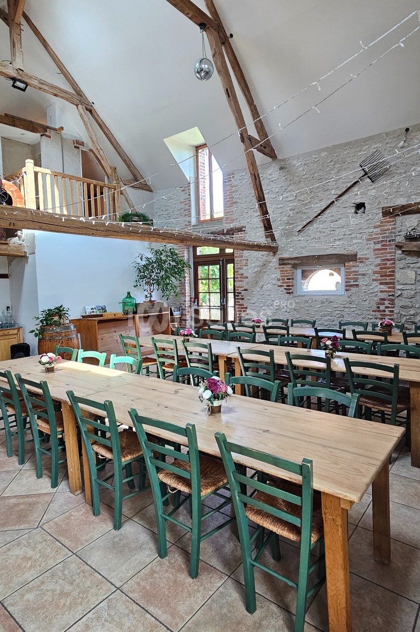 Salle lumineuse avec de grandes tables en bois, chaises vertes, poutres apparentes et murs en pierre.