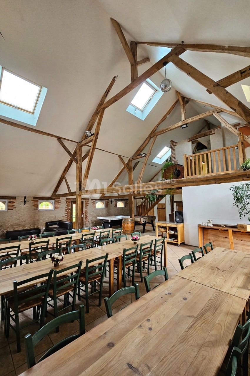 Salle lumineuse avec poutres apparentes, grandes tables en bois, chaises vertes et baies vitrées laissant entrer la lumière.