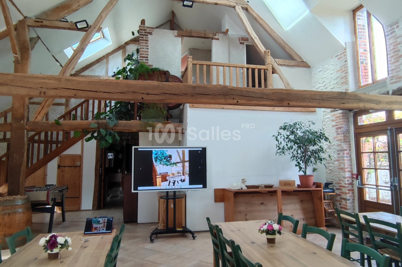 Salle lumineuse avec poutres apparentes, tables en bois, plantes, écran de présentation et billard en arrière-plan.