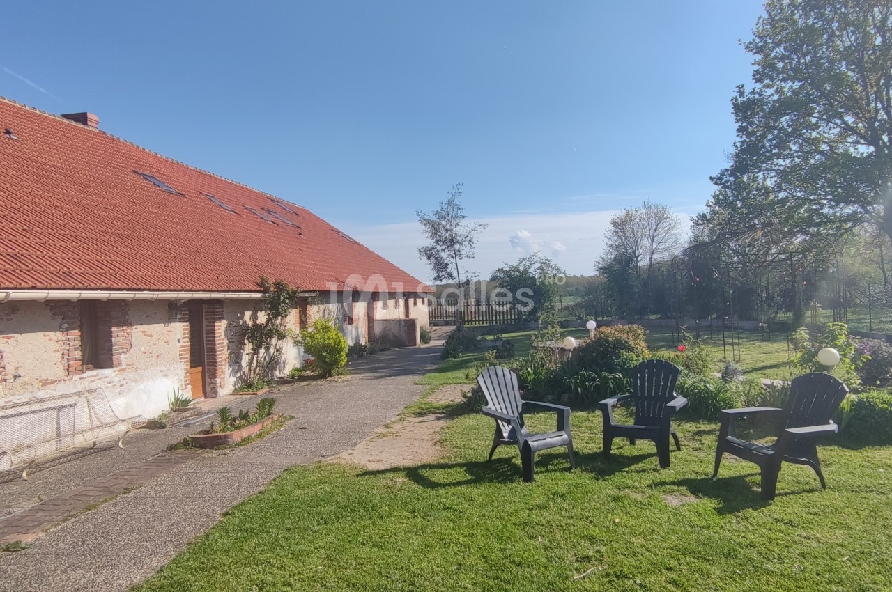 Cour d'une maison en briques avec toit en tuiles rouges, jardin aménagé et chaises disposées sur une pelouse.