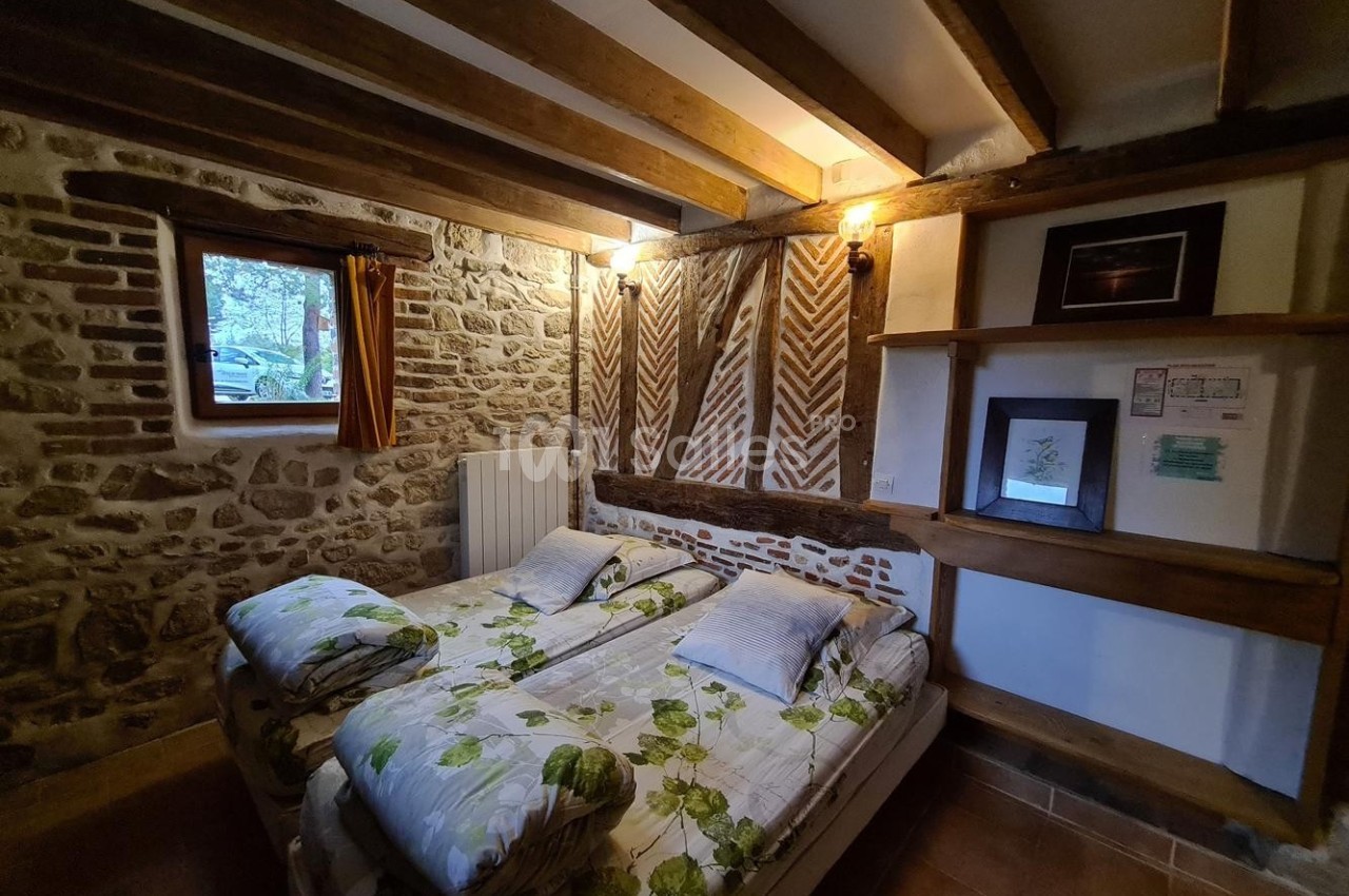 Chambre rustique avec murs en pierre, poutres apparentes, lit double et petite fenêtre donnant sur l'extérieur.