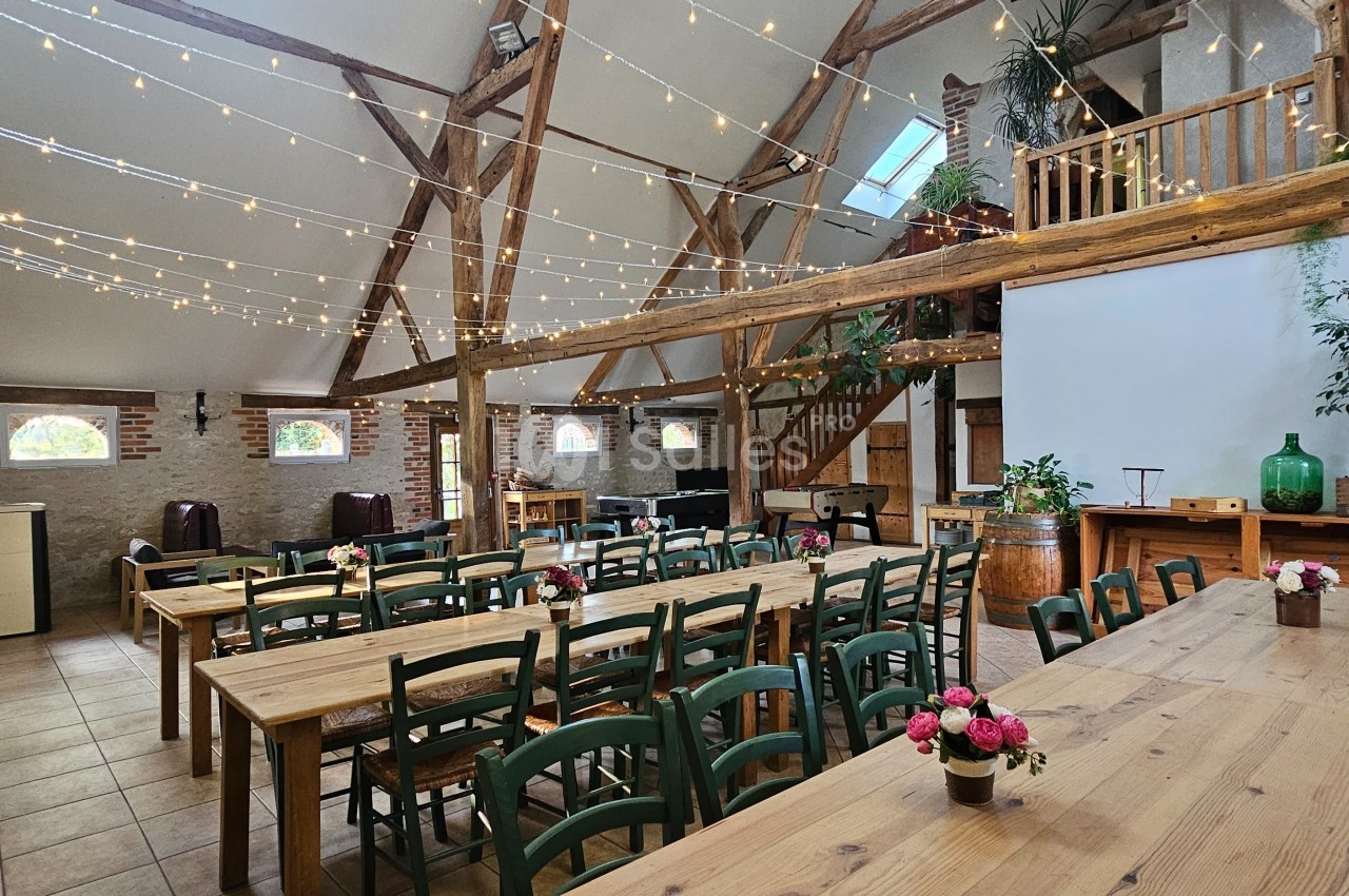 Salle lumineuse avec poutres apparentes, tables en bois, chaises vertes, guirlandes lumineuses et décorations florales.