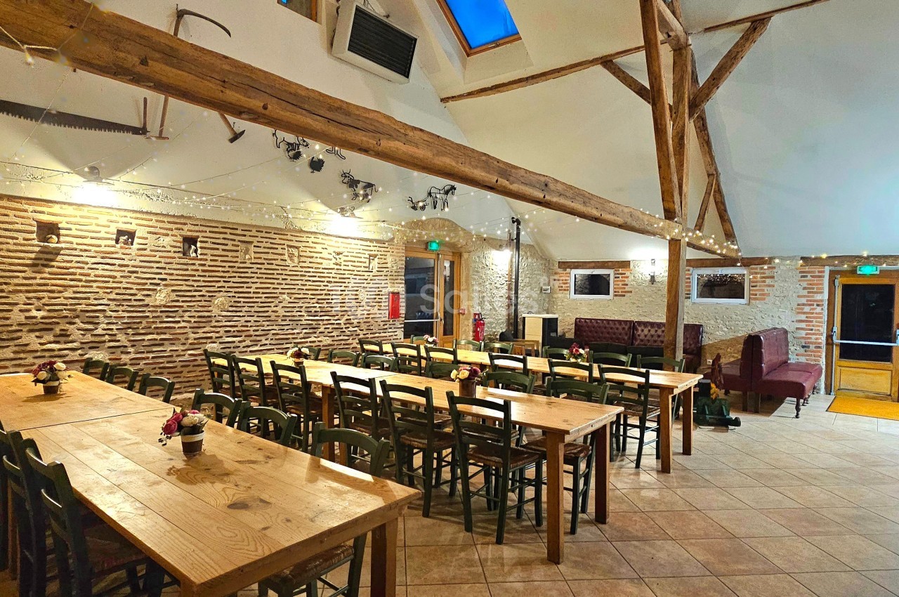 Salle lumineuse avec murs en briques, poutres apparentes, tables en bois et chaises alignées pour un événement.