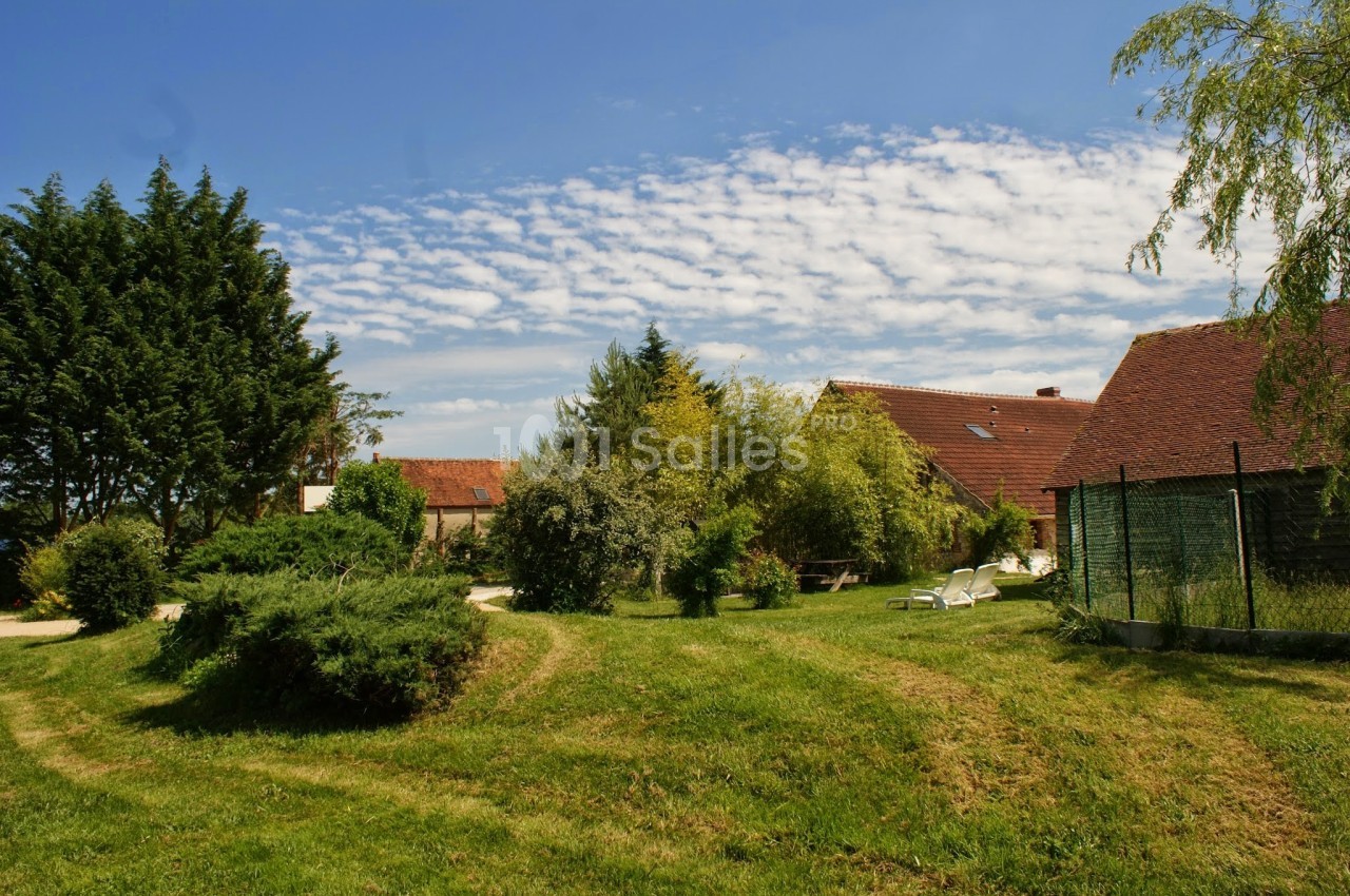 Jardin verdoyant avec pelouse, arbres, buissons et maisons en arrière-plan sous un ciel bleu parsemé de nuages.