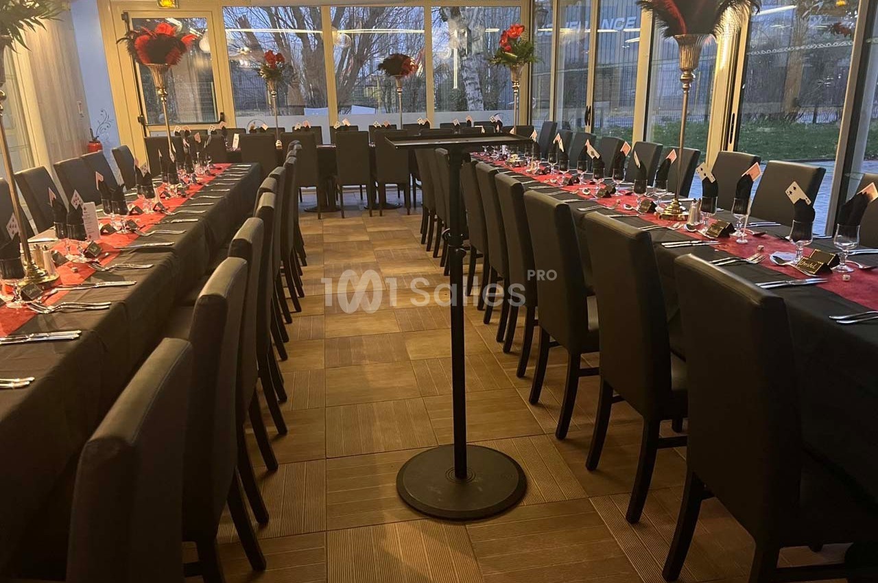 Salle de réception avec deux longues tables dressées, nappes rouges, chaises noires et grandes baies vitrées en arrière-plan.