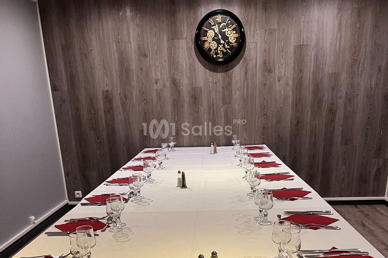 Salle à manger avec une grande table dressée, nappée de blanc, couverts alignés et horloge murale en arrière-plan.