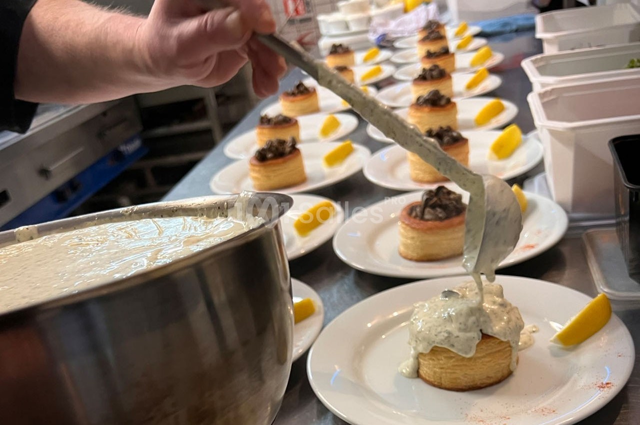 Un chef verse une sauce blanche sur des bouchées garnies, alignées sur des assiettes dans une cuisine professionnelle.