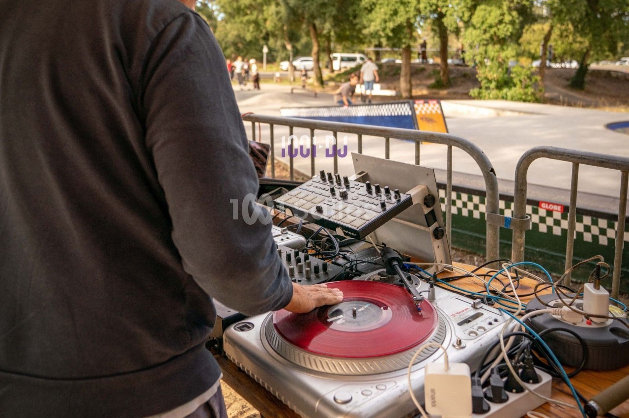 Un DJ manipule un disque vinyle rouge sur une platine, entouré d'équipements audio en extérieur près d'un skatepark.
