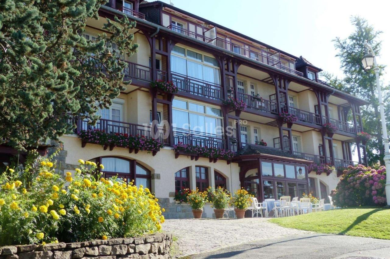 Façade d'un hôtel en bois et pierre avec balcons fleuris, entouré de fleurs et d'arbres dans un cadre ensoleillé.