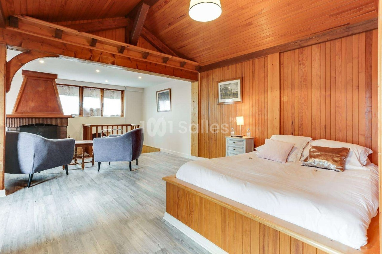 Chambre lumineuse avec lit double, murs en bois, plafond en poutres apparentes, coin salon et cheminée.