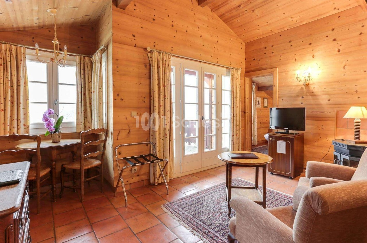Salon chaleureux avec murs en bois, table près d'une fenêtre, fauteuils, télévision et porte-fenêtre donnant sur l'extérieur.