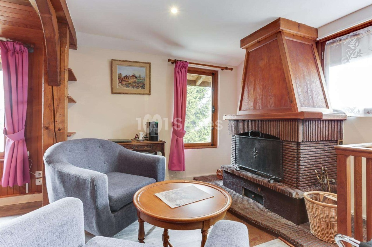 Salon chaleureux avec cheminée en briques, fauteuils gris, table basse en bois et rideaux roses près des fenêtres.