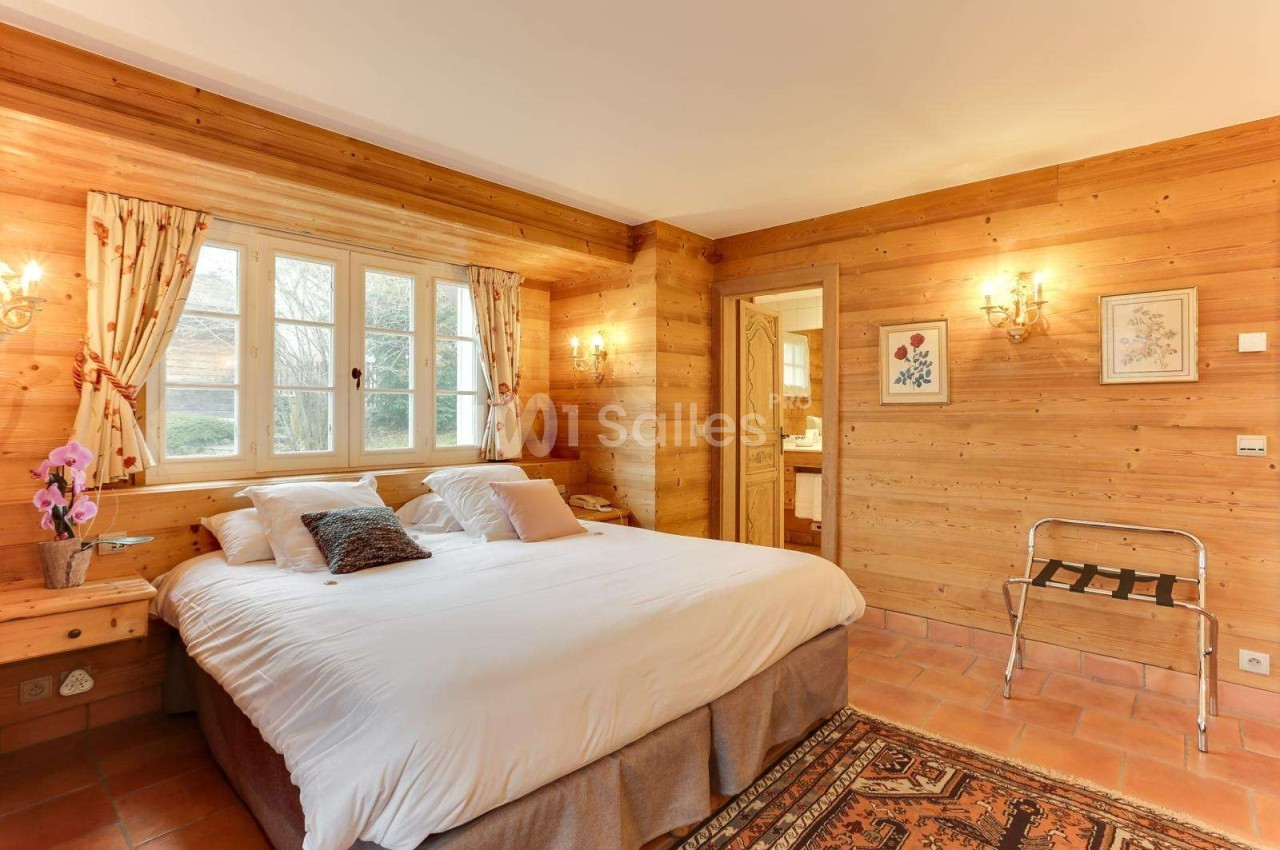 Chambre avec murs en bois, lit double, fenêtres avec rideaux fleuris, éclairage mural et sol en carreaux.