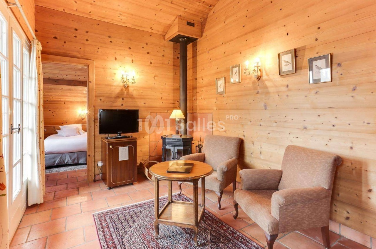 Salon chaleureux avec murs en bois, poêle à bois, deux fauteuils, table basse, tapis et vue sur une chambre adjacente.