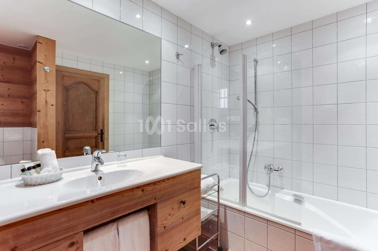 Salle de bain avec carrelage blanc, baignoire-douche, meuble en bois avec lavabo et miroir éclairé.