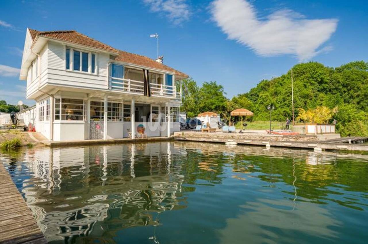 Maison blanche à deux étages au bord d'un plan d'eau, entourée de végétation sous un ciel bleu.