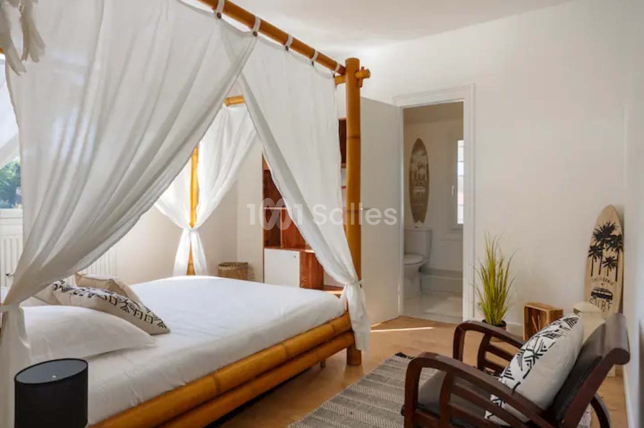 Chambre lumineuse avec lit à baldaquin en bois, fauteuils, décoration tropicale et accès à une salle de bain.