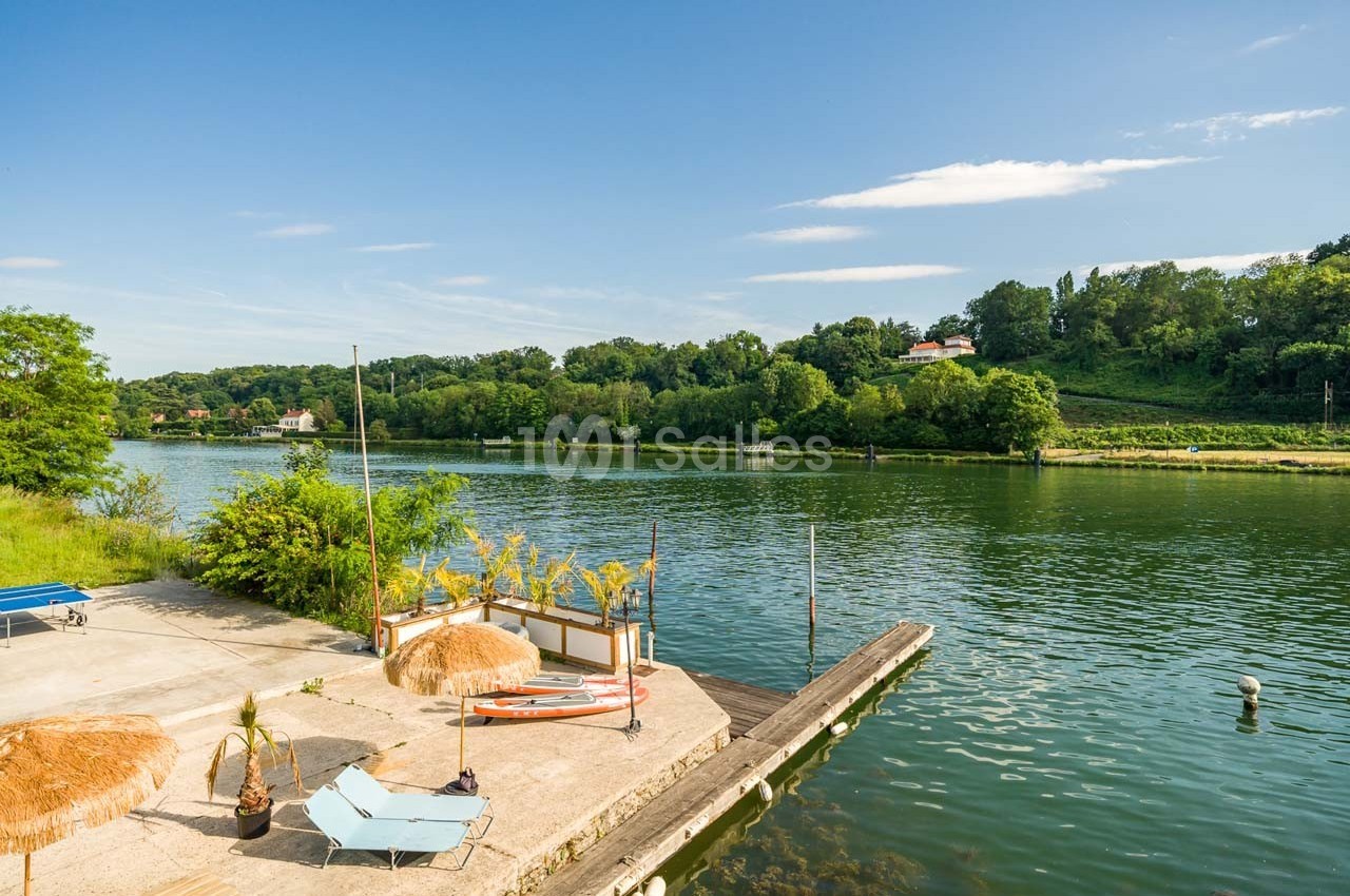 Terrasse avec transats, parasols et ponton en bord de rivière, entourée de verdure sous un ciel dégagé.