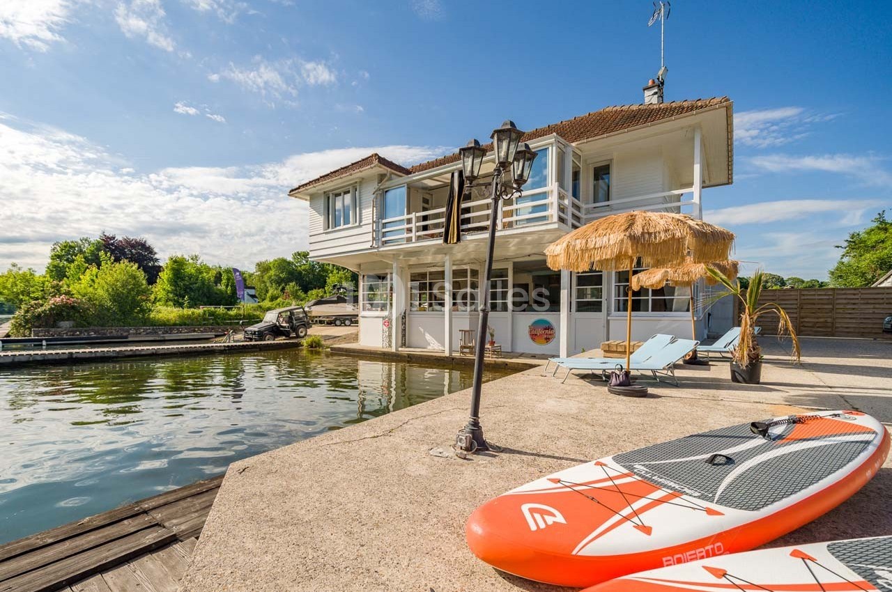 Maison au bord de l'eau avec terrasse, transats, parasol en paille et planche de paddle sur un quai ensoleillé.