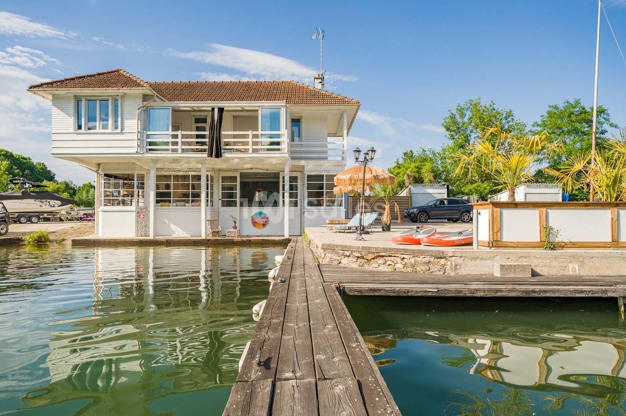Maison blanche sur pilotis au bord d'un canal, avec terrasse aménagée, kayaks et végétation environnante.