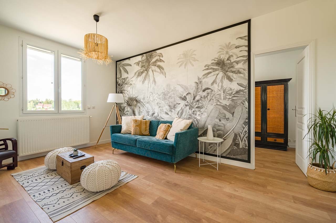 Salon lumineux avec canapé vert, poufs en osier, table basse en bois et mur décoré d'une fresque tropicale en noir et blanc.