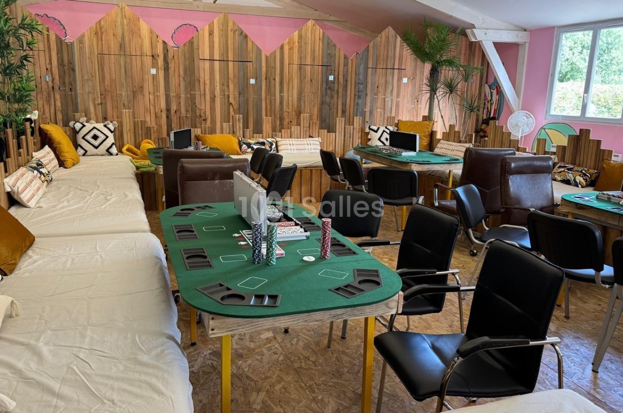 Salle aménagée avec des tables de jeu de cartes, des chaises, des canapés et une décoration chaleureuse en bois et plantes.