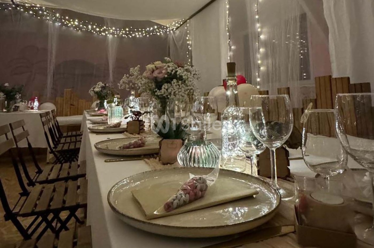 Table décorée pour un dîner festif avec vaisselle élégante, fleurs, bougies et guirlandes lumineuses en arrière-plan.