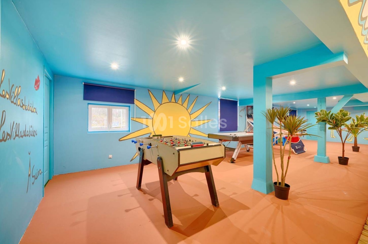 Salle de jeux lumineuse avec baby-foot, billard, murs bleus décorés d'un soleil et plantes en pots.