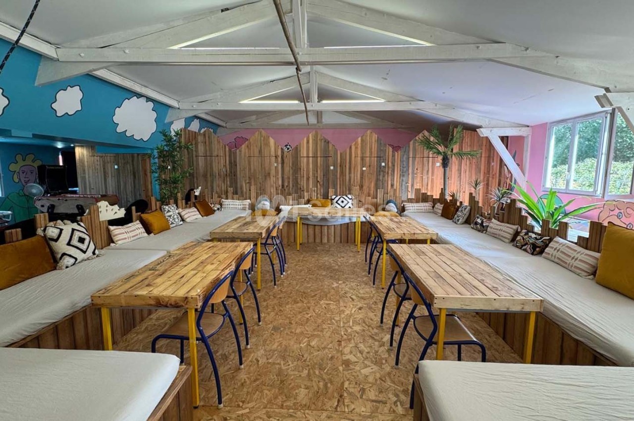 Salle lumineuse avec tables en bois, bancs garnis de coussins et décorations murales colorées.