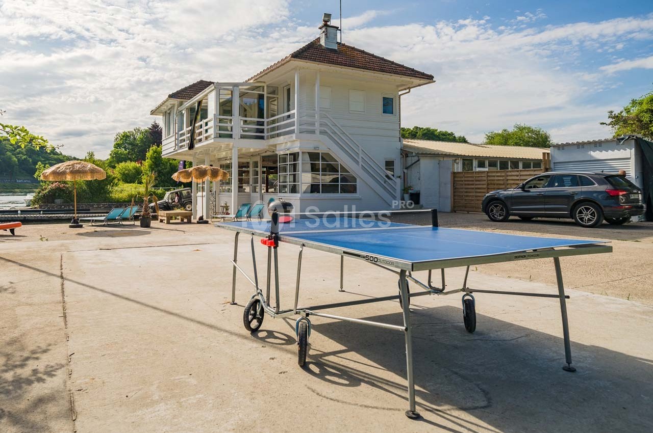 Table de ping-pong sur une terrasse ensoleillée devant une maison blanche à deux étages, avec parasols et voiture garée.