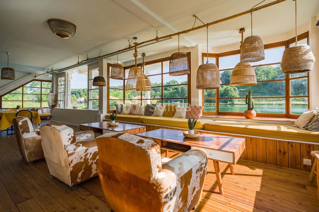 Salle lumineuse avec grandes fenêtres, mobilier en bois, fauteuils en tissu et suspensions en osier.