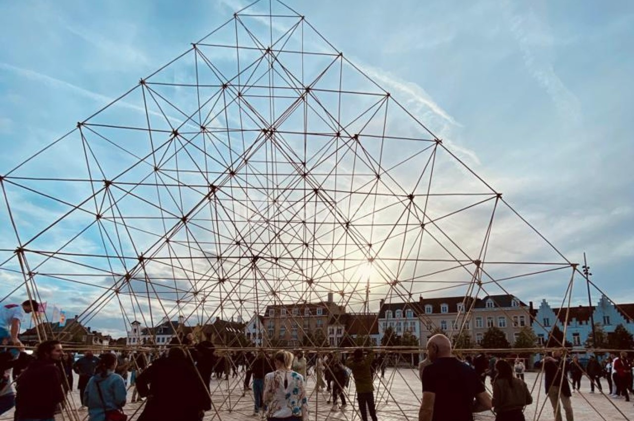 Structure géométrique métallique sur une place publique, entourée de passants sous un ciel partiellement nuageux.