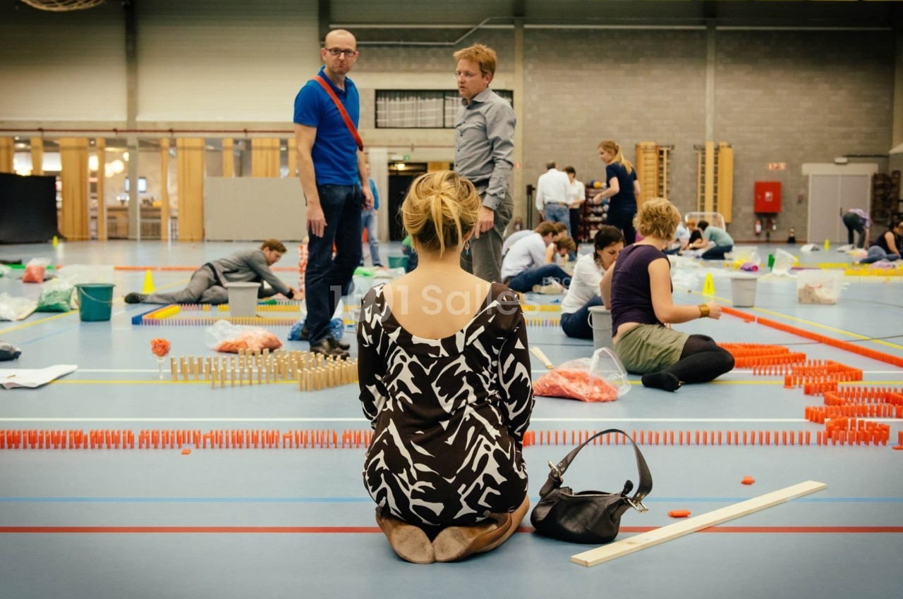 Des personnes assemblent des dominos colorés dans un gymnase, une femme assise au premier plan les observe.