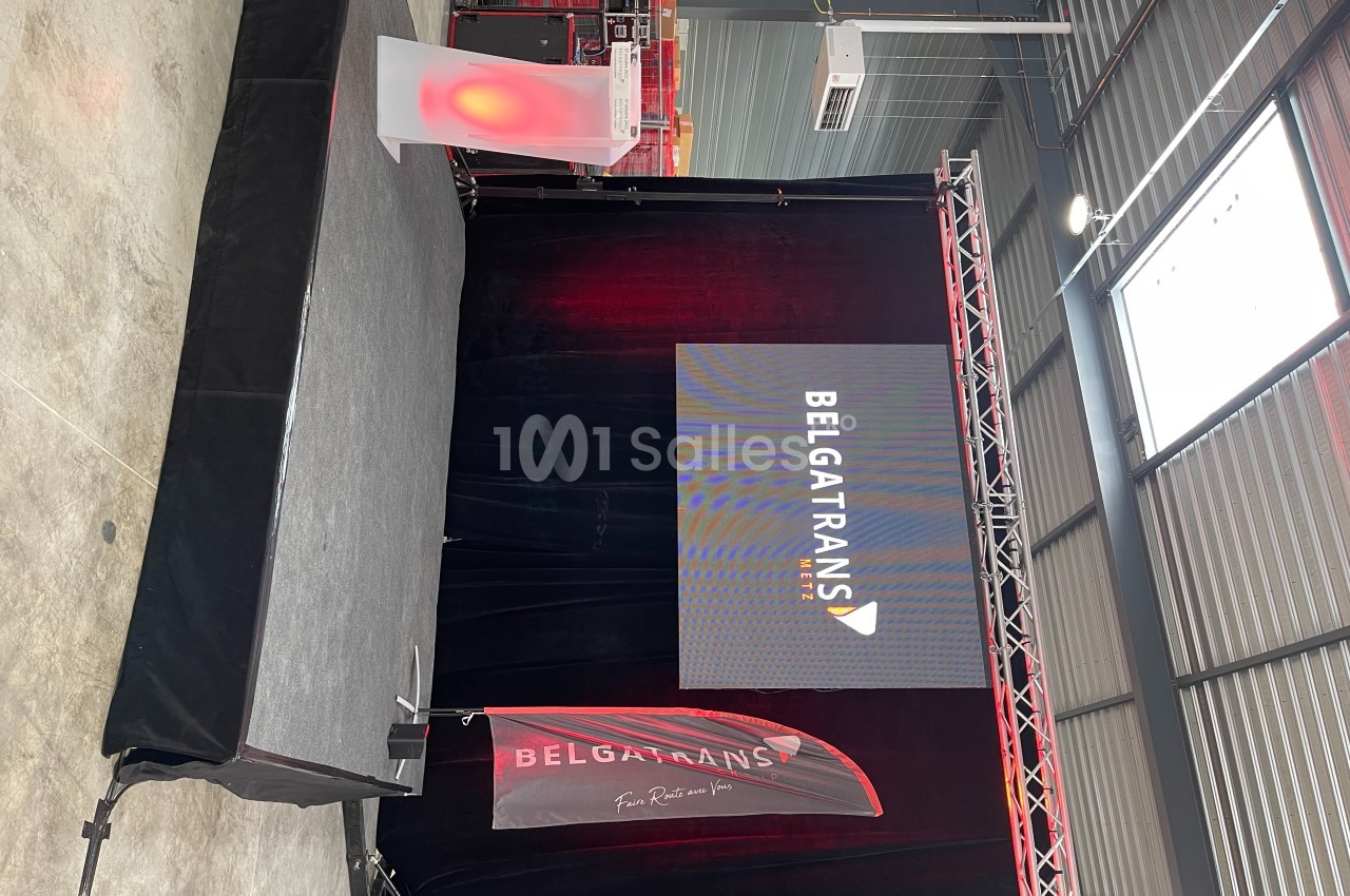 Scène avec écran affichant ’Belga Trans’, éclairage rouge, pupitre lumineux et drapeau dans un espace industriel.
