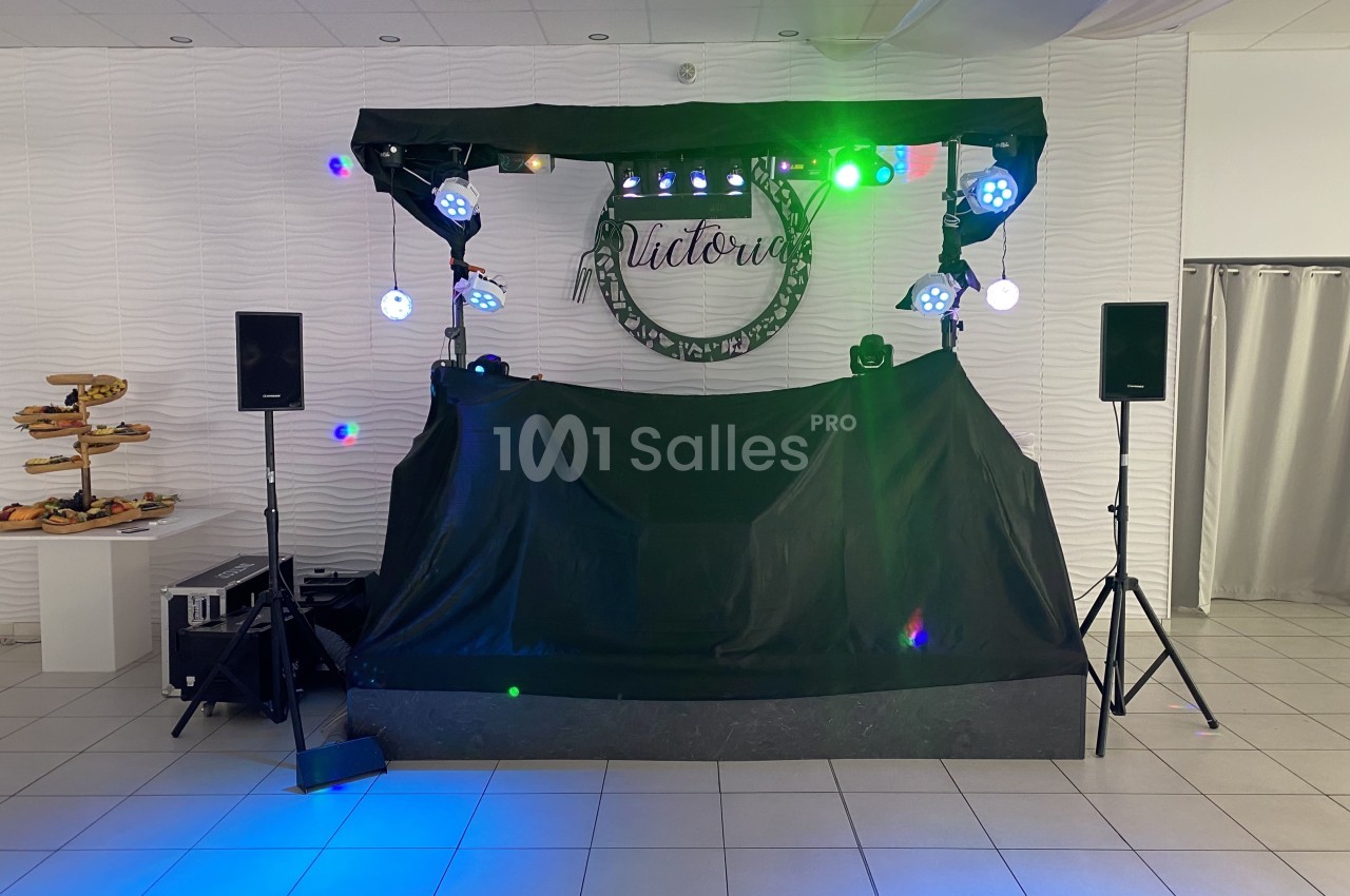 Scène de DJ avec éclairage coloré, deux enceintes et une table recouverte, dans une salle au décor sobre.