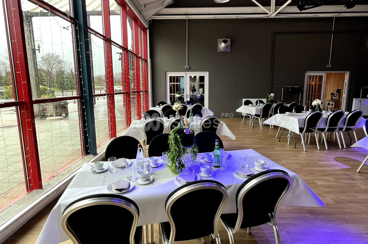 Salle lumineuse avec grandes baies vitrées, tables dressées avec nappes blanches, vaisselle et décorations végétales.