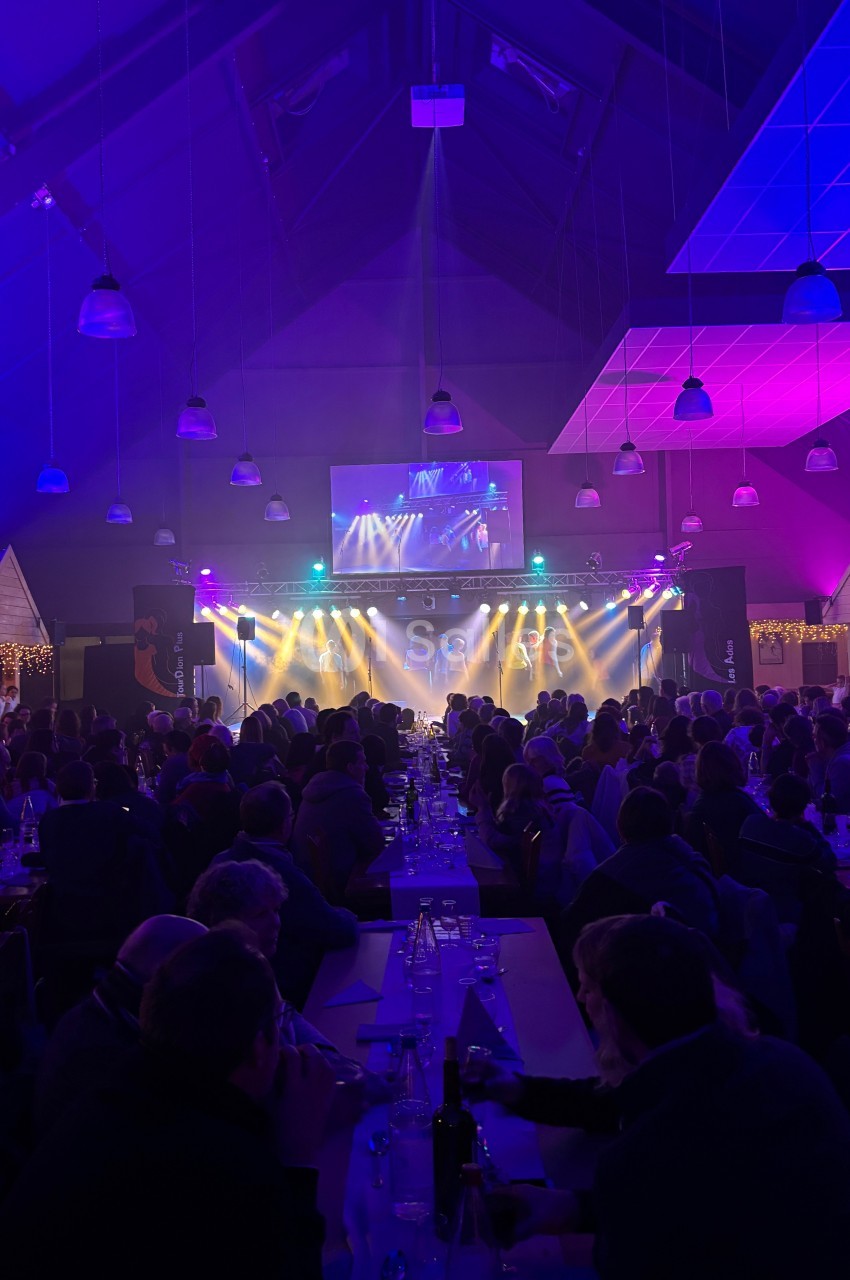 Salle comble avec des tables alignées, éclairage coloré et scène illuminée au fond diffusant un spectacle.