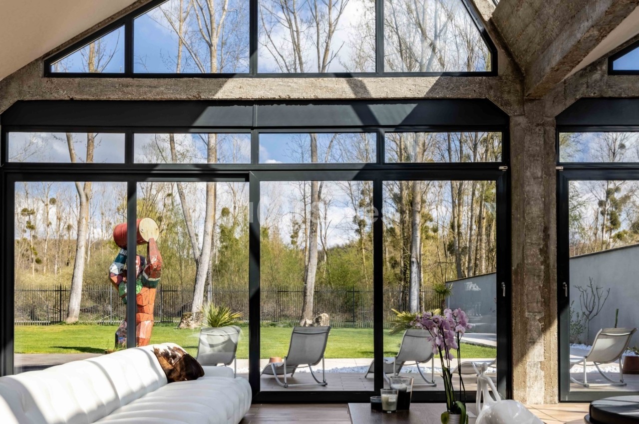 Salon lumineux avec grande baie vitrée donnant sur un jardin, canapé blanc au premier plan et orchidée sur la table.