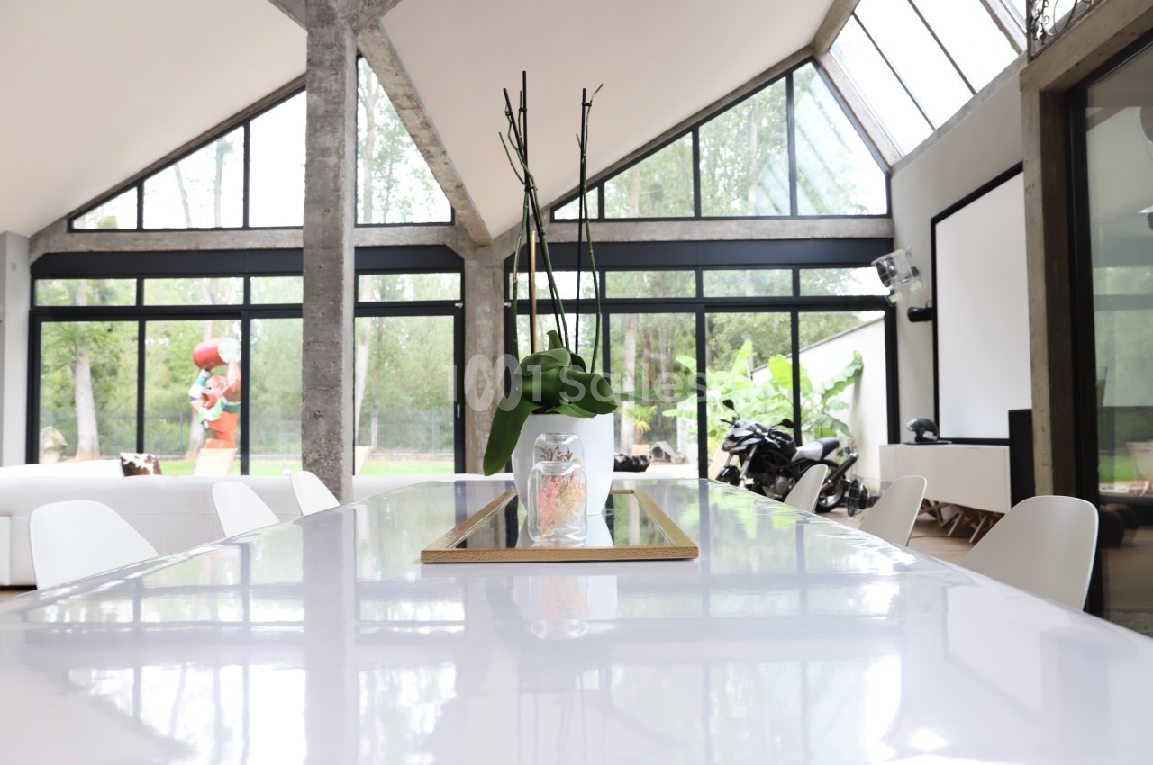 Table blanche avec un vase contenant une plante, dans une pièce lumineuse aux grandes baies vitrées donnant sur un jardin.