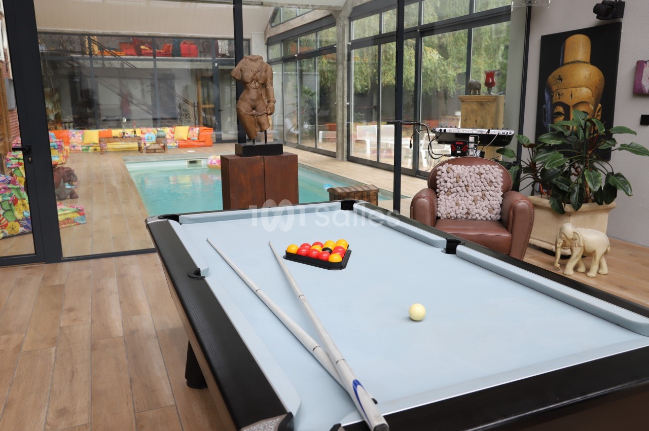 Table de billard avec accessoires au premier plan, piscine intérieure et espace de jeux visibles en arrière-plan.